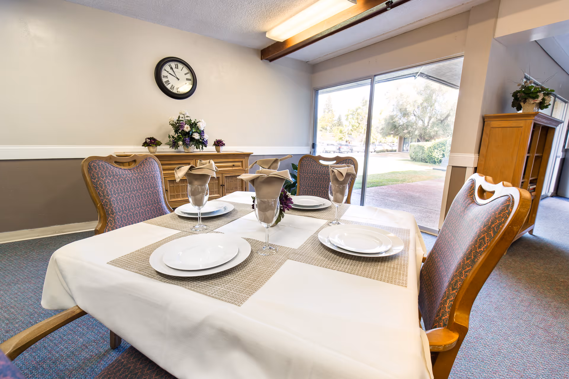 A dining room table set for four with white plates, glasses with beige napkins, and placemats. The room has upholstered chairs, a sideboard with flower arrangements, a wall clock, and large sliding glass doors showing an outdoor view with trees and a walkway.