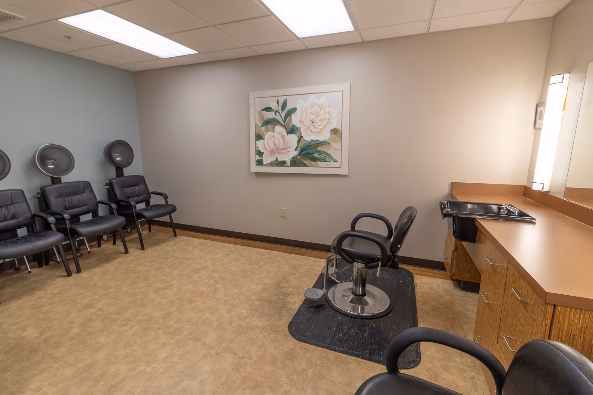 Interior salon room with multiple waiting chairs and hooded dryers, a styling chair by a counter and sink, and floral wall art.