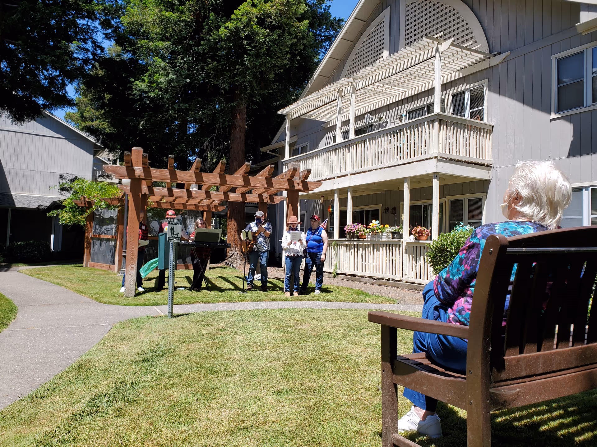 An elderly woman sits on a bench watching a small musical group perform under a wooden pergola in the courtyard of a multi-story senior living building.