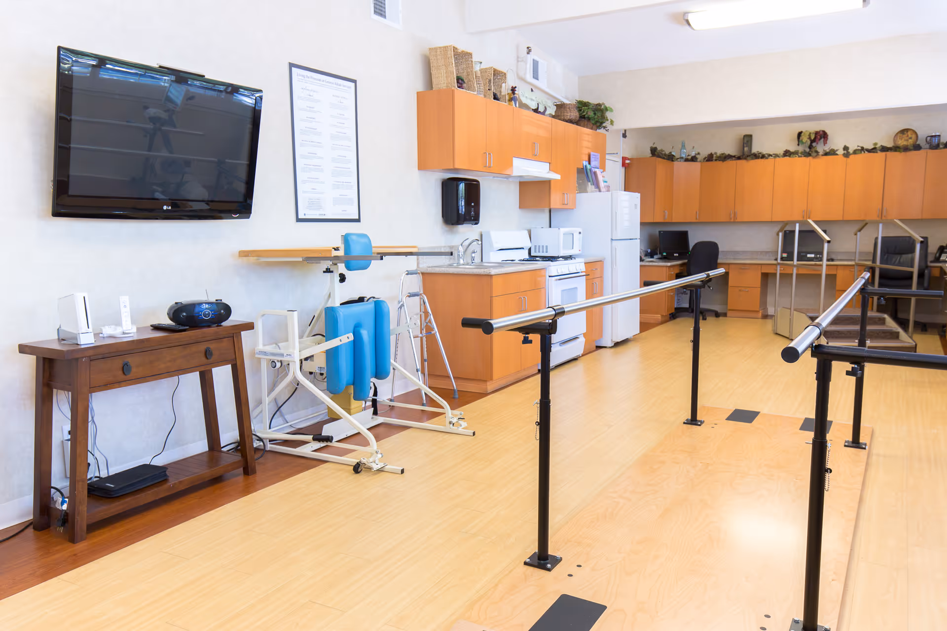 A rehabilitation or physical therapy room with parallel bars for walking exercises, a blue padded exercise machine, a walker, a wooden table with electronic devices, a wall-mounted TV, and a kitchenette area with wooden cabinets, a stove, microwave, and refrigerator.