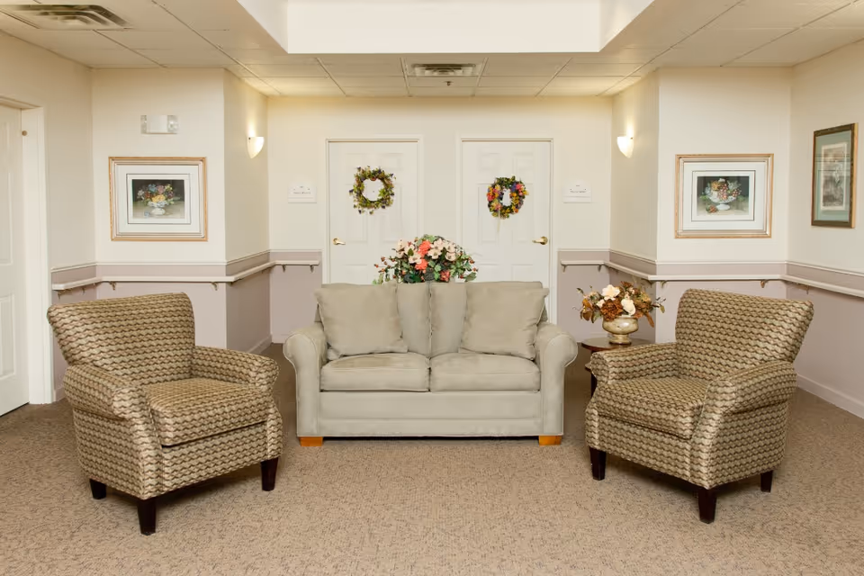Seating area with a beige loveseat flanked by two patterned armchairs in a decorated hallway/lounge with floral arrangements and framed art.