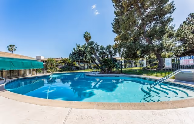 Outdoor swimming pool with clear blue water surrounded by a concrete deck, green trees, and a fence. There is a green awning on the left side and a warning sign on the right side near the pool steps. The sky is clear with a few clouds.
