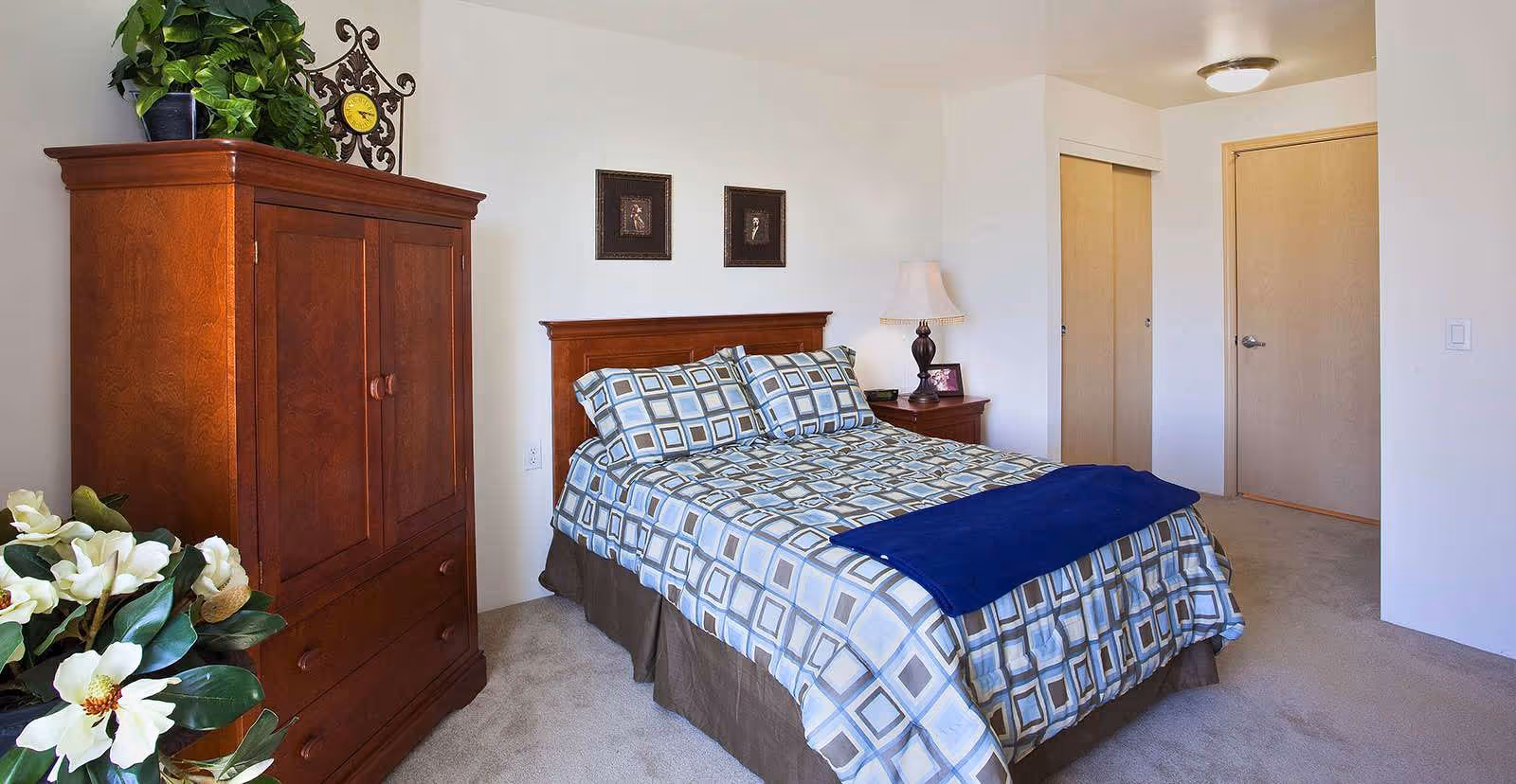 A neatly made bed with a geometric patterned bedspread and two matching pillows in a bedroom. To the left of the bed is a wooden armoire with a plant and decorative clock on top. A nightstand with a lamp and framed photo is beside the bed. The room has beige carpet and white walls with two framed pictures above the bed. There are two closed wooden doors on the right side of the room.