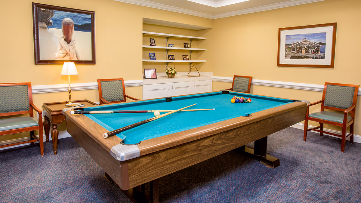 A recreational room with a pool table in the center, four wooden chairs with green cushions arranged around the room, a small side table with a lamp, and framed pictures on the yellow walls. There are shelves with small decorative items and a carpeted floor.
