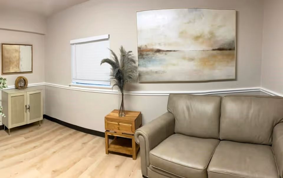 Interior seating area with a beige leather couch, wooden side table holding a vase of tall pampas grass, a small cabinet, window, and wall art.