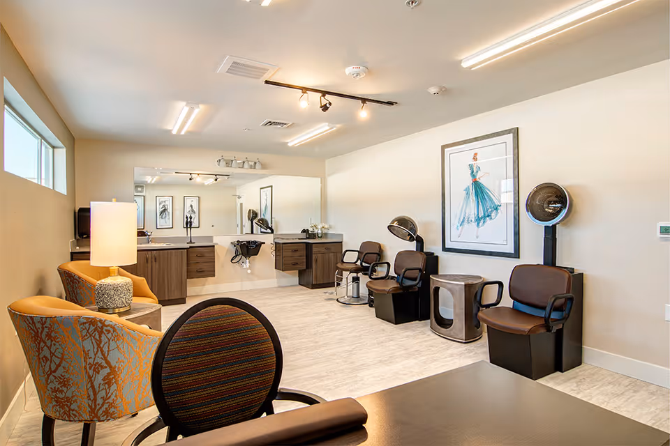 A bright and clean salon area in a senior living community featuring three brown salon chairs with hair dryers, a large mirror above two wooden cabinets with sinks, a small table, and two patterned armchairs with a lamp near a window.