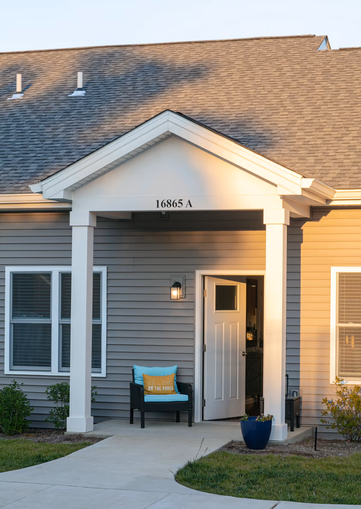 Entrance of a residential building with the address number 16865 A above the porch. The porch has a black chair with a blue cushion and a yellow pillow that says 'Life is better ON THE PORCH'. The door is open, and there is a blue planter with flowers near the entrance. The building has gray siding and white trim.