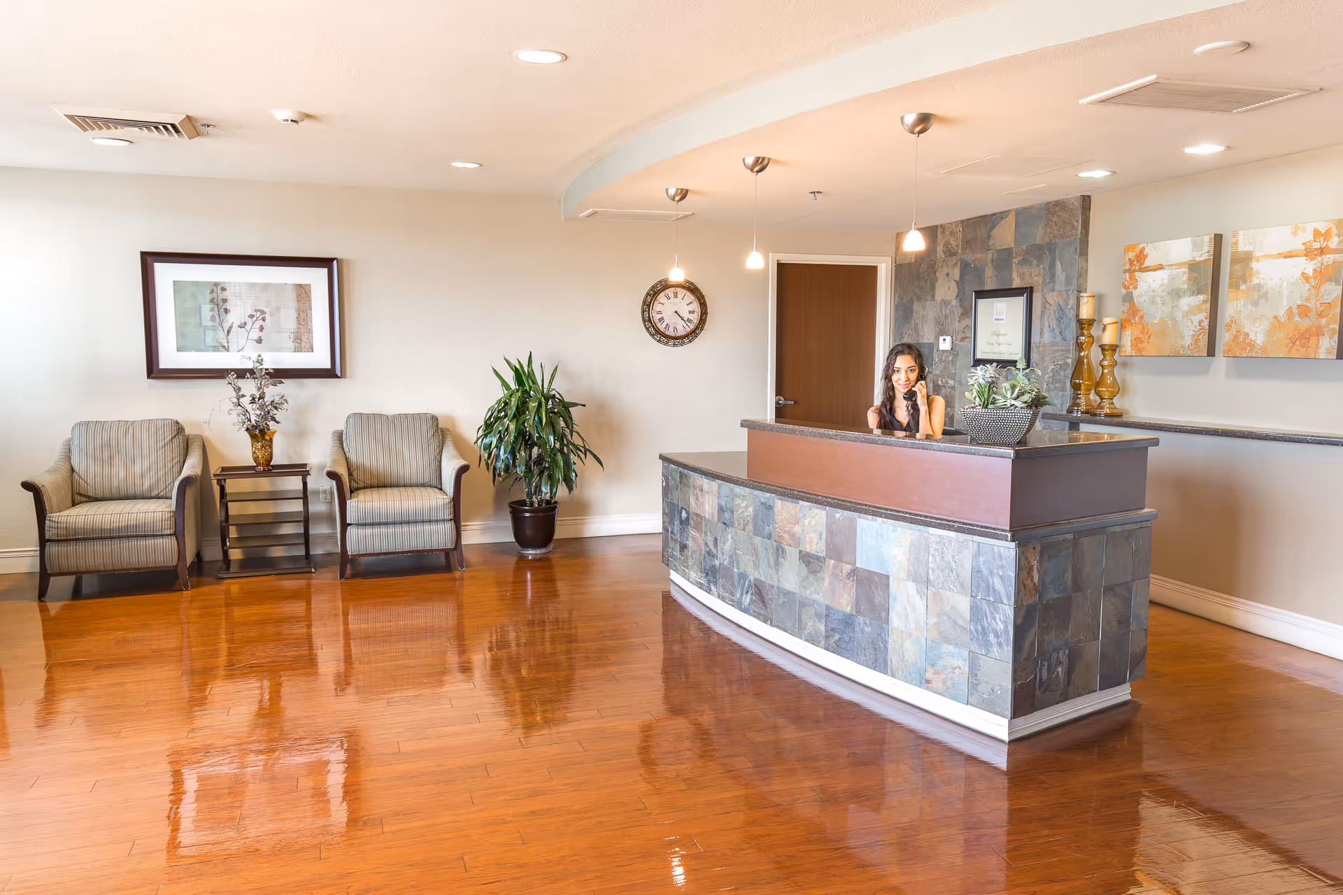 Reception area with a curved stone and wood front desk where a woman is seated, a wall clock, two armchairs with a small table and a plant between them, and decorative artwork on the walls. The floor is polished wood.