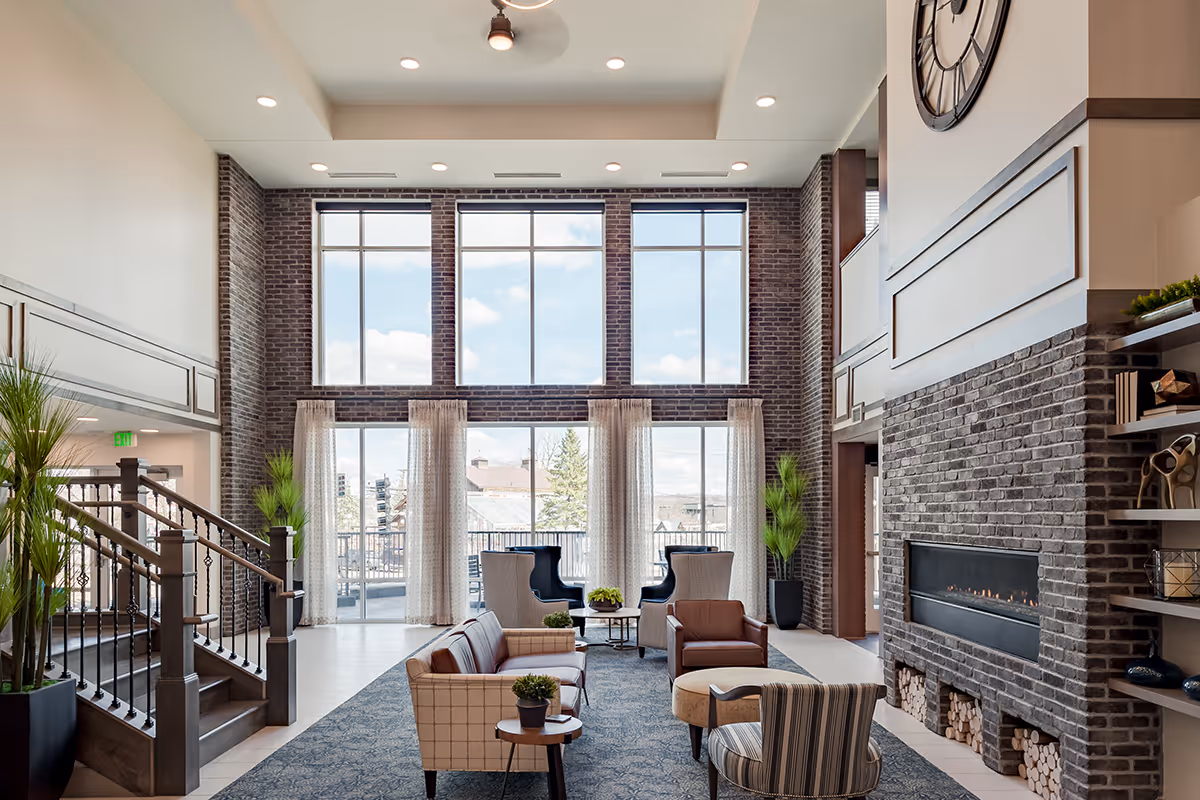 A spacious and well-lit living room area with large floor-to-ceiling windows letting in natural light. The room features a modern brick fireplace with stacked firewood underneath, several comfortable chairs and sofas arranged around small tables, decorative plants, and a staircase with wooden railings on the left side. The ceiling has recessed lighting and a ceiling fan, and a large clock is mounted above the fireplace.