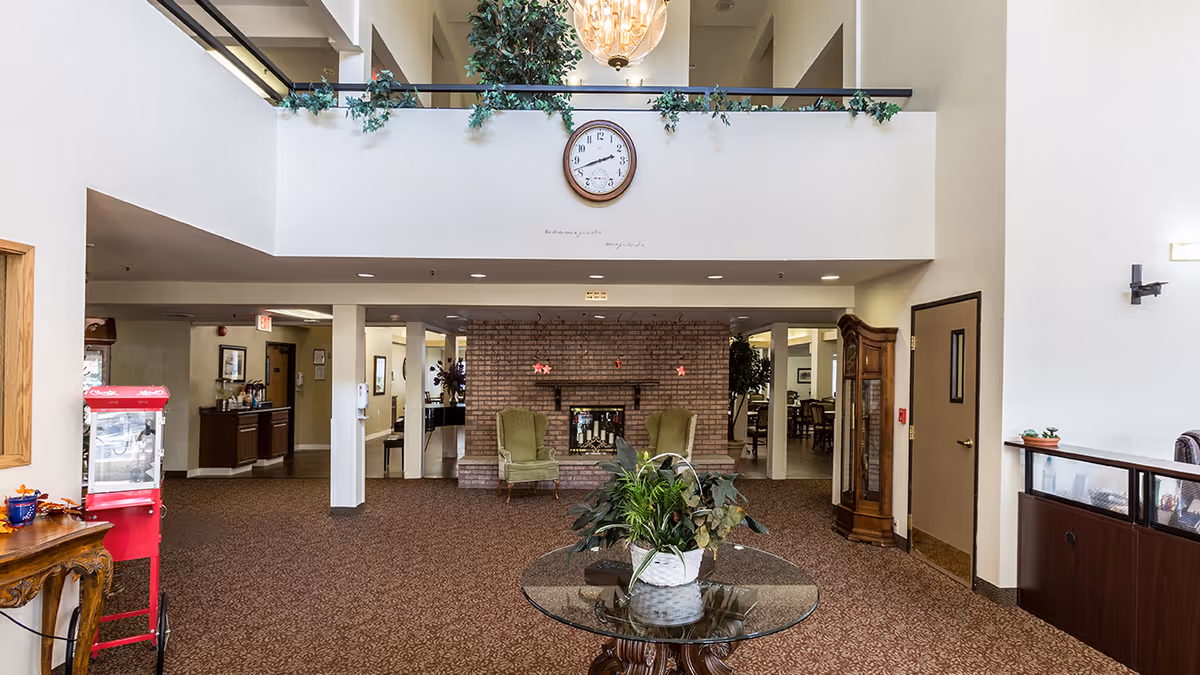 Spacious senior living lobby with a central glass table and plant, seating in front of a brick fireplace, a clock above and a chandelier on the balcony level.