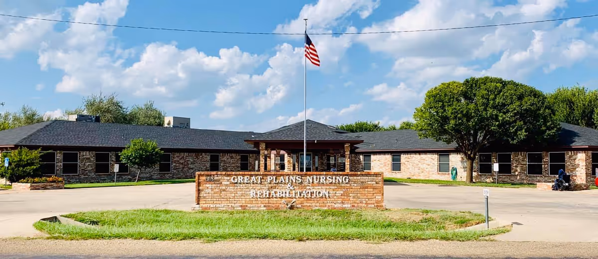 Exterior view of Great Plains Nursing and Rehabilitation facility with a brick sign in front displaying the facility name. The building is single-story with a dark roof, surrounded by trees and greenery under a partly cloudy blue sky. An American flag flies on a flagpole behind the sign.