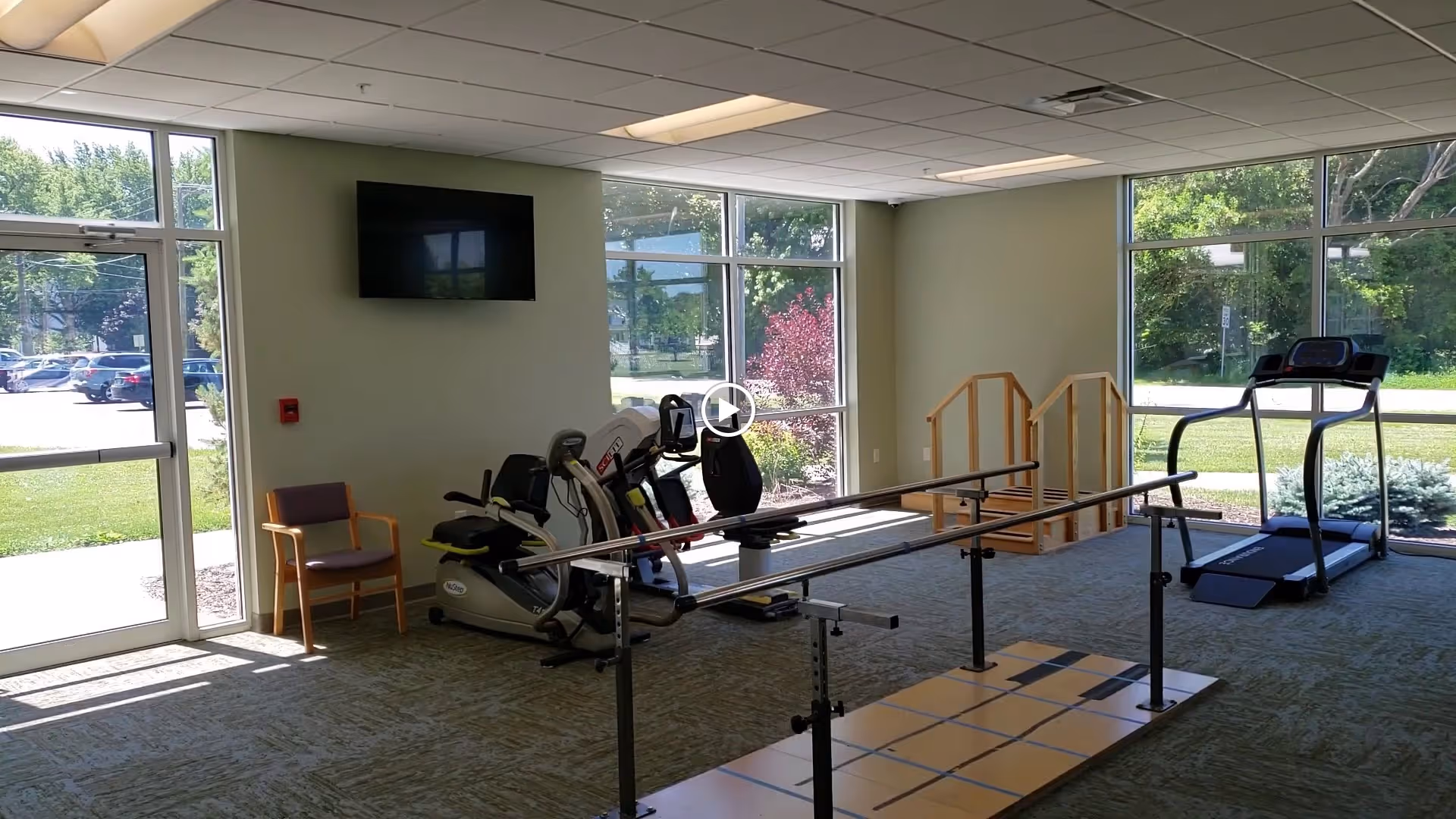 A rehabilitation exercise room with large windows letting in natural light. The room contains parallel bars for walking exercises, a treadmill, a recumbent exercise bike, a small set of wooden stairs for step training, a wall-mounted TV, and a single chair near the entrance door. Outside the windows, greenery and parked cars are visible.