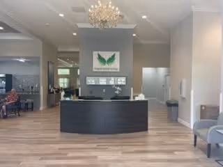 Reception area of Angels Senior Living At The Reserves Of Idlewild featuring a modern black reception desk with a decorative plant and tissue box on top. Behind the desk is a wall with a framed artwork of green leaves and the facility name. The space has light wood flooring, neutral-colored walls, a chandelier, and seating areas visible on both sides.