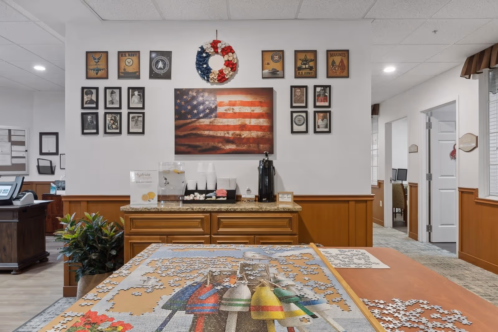 Interior view of a senior living facility room with a partially completed jigsaw puzzle on a table in the foreground. The back wall features a large American flag artwork, a red, white, and blue wreath, and framed military-themed pictures and portraits. Below the wall art is a countertop with a water dispenser, cups, napkins, and a coffee station. The room has wood paneling on the lower half of the walls and white walls above, with doorways leading to other rooms visible on the right side.