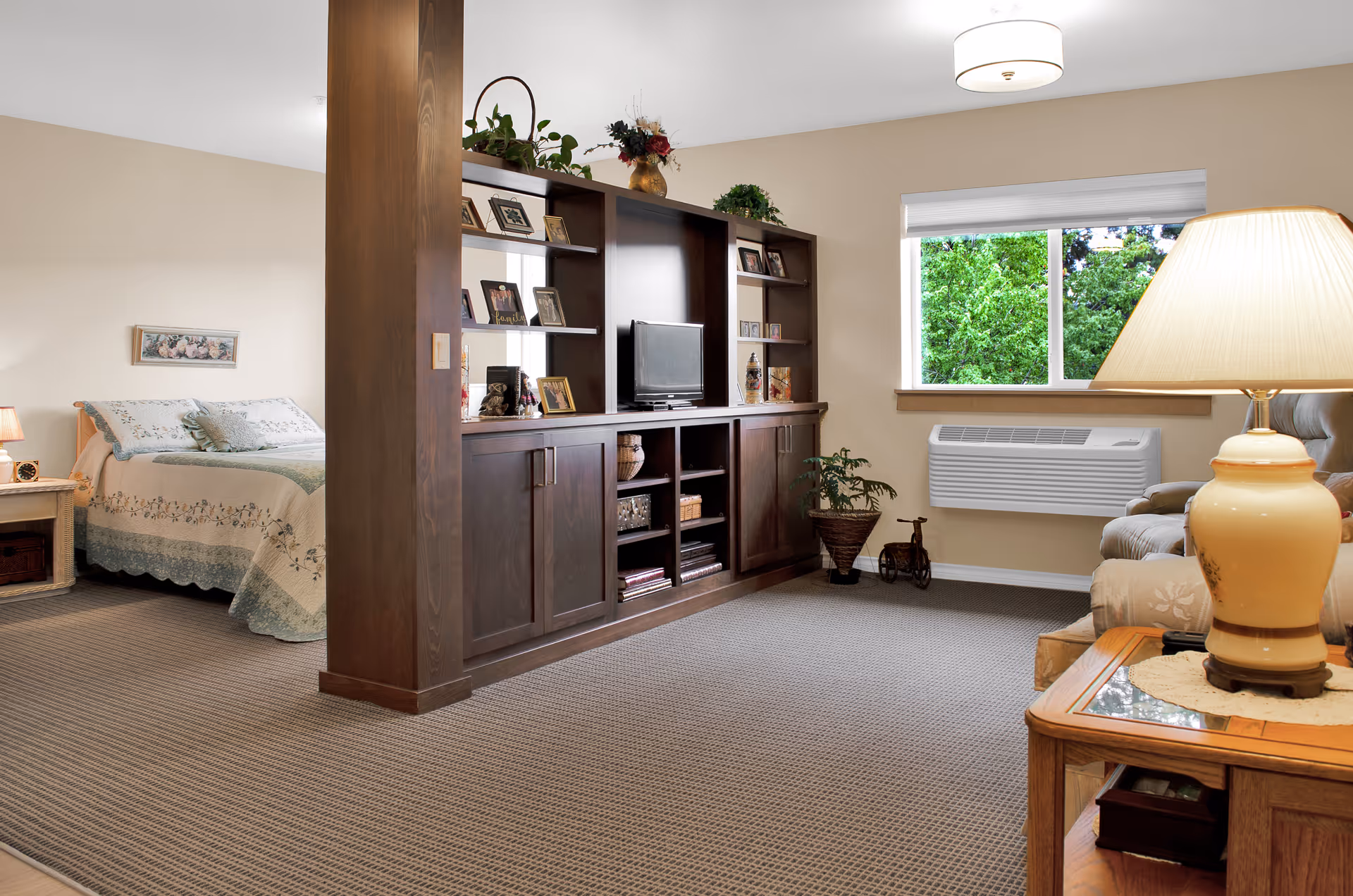 A cozy senior living studio interior with a bed on the left, wooden entertainment shelves and a TV in the center, and a seating area with a lamp by a window on the right.