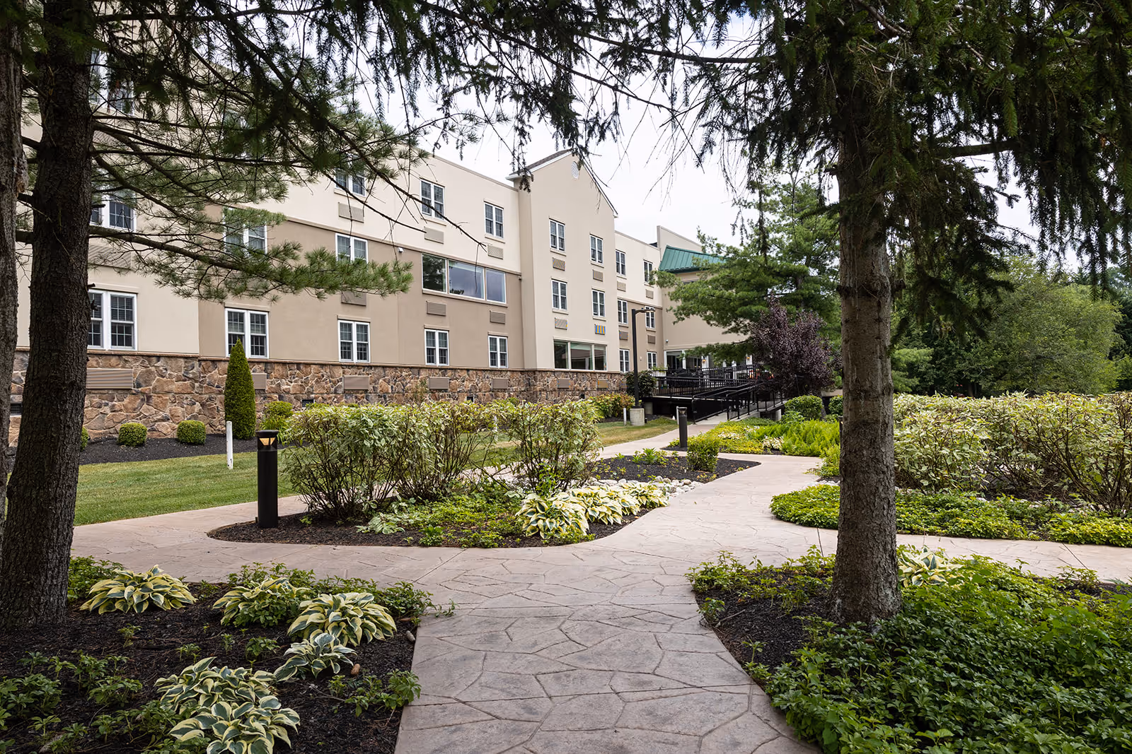 View of the exterior of a multi-story senior living facility building with beige and stone facade, surrounded by well-maintained landscaped gardens and paved walkways, framed by trees in the foreground.