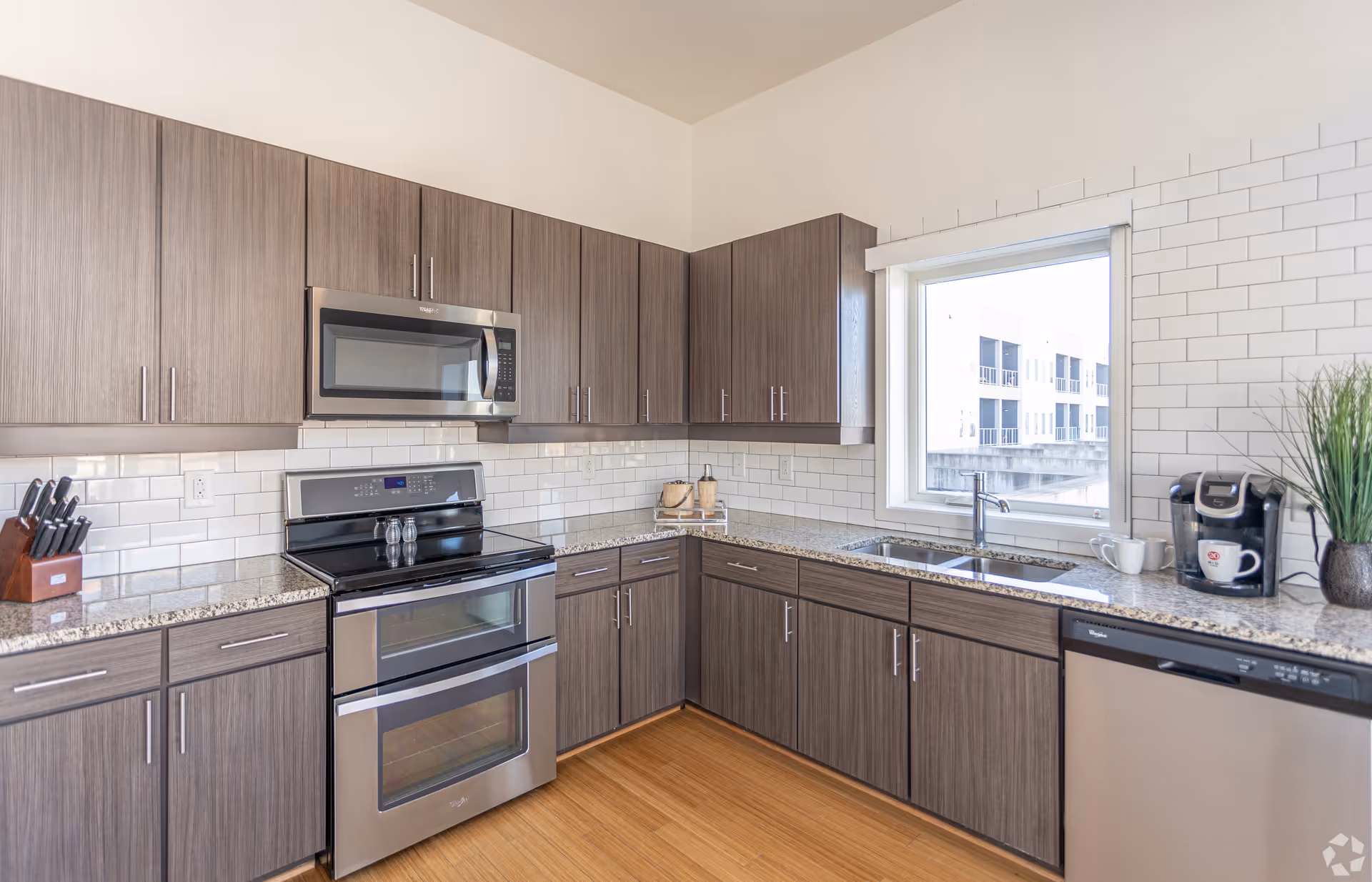 Modern kitchen with stainless steel appliances including a double oven and microwave, dark wood cabinets, granite countertops, a double sink under a window, a coffee maker, and a knife block on the counter. The backsplash is white subway tile and the floor is wood.