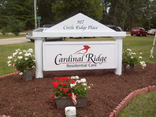 Outdoor sign for Cardinal Ridge Residential Care located at 817 Circle Ridge Place, surrounded by flower beds with red and white flowers and a grassy area with trees and parked cars in the background.