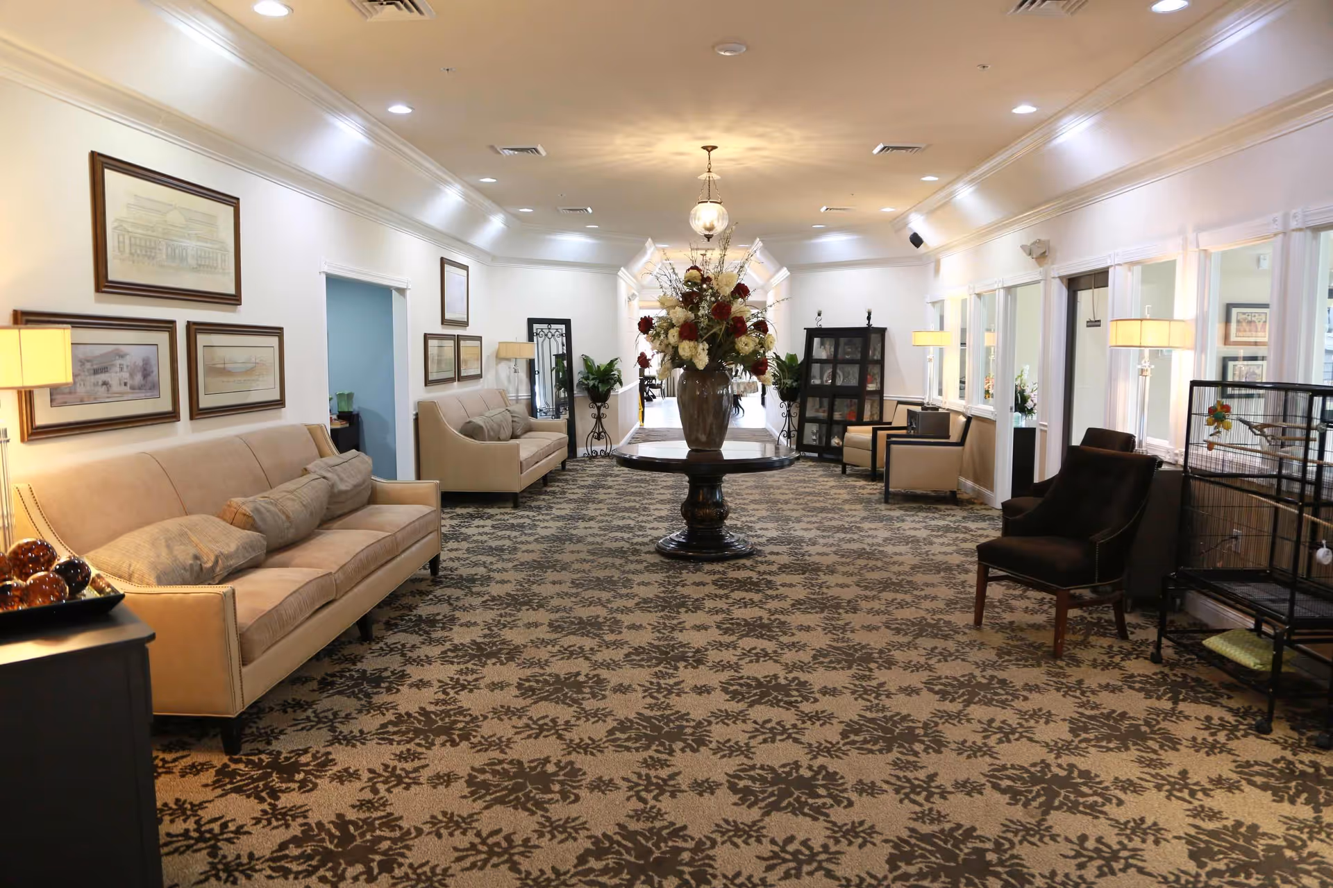 A spacious, well-lit lounge area with beige sofas and armchairs arranged along the walls. A round table with a large floral arrangement is centered on a patterned carpet. The walls are decorated with framed artwork, and there are several lamps providing warm lighting. The room has a welcoming and comfortable atmosphere.