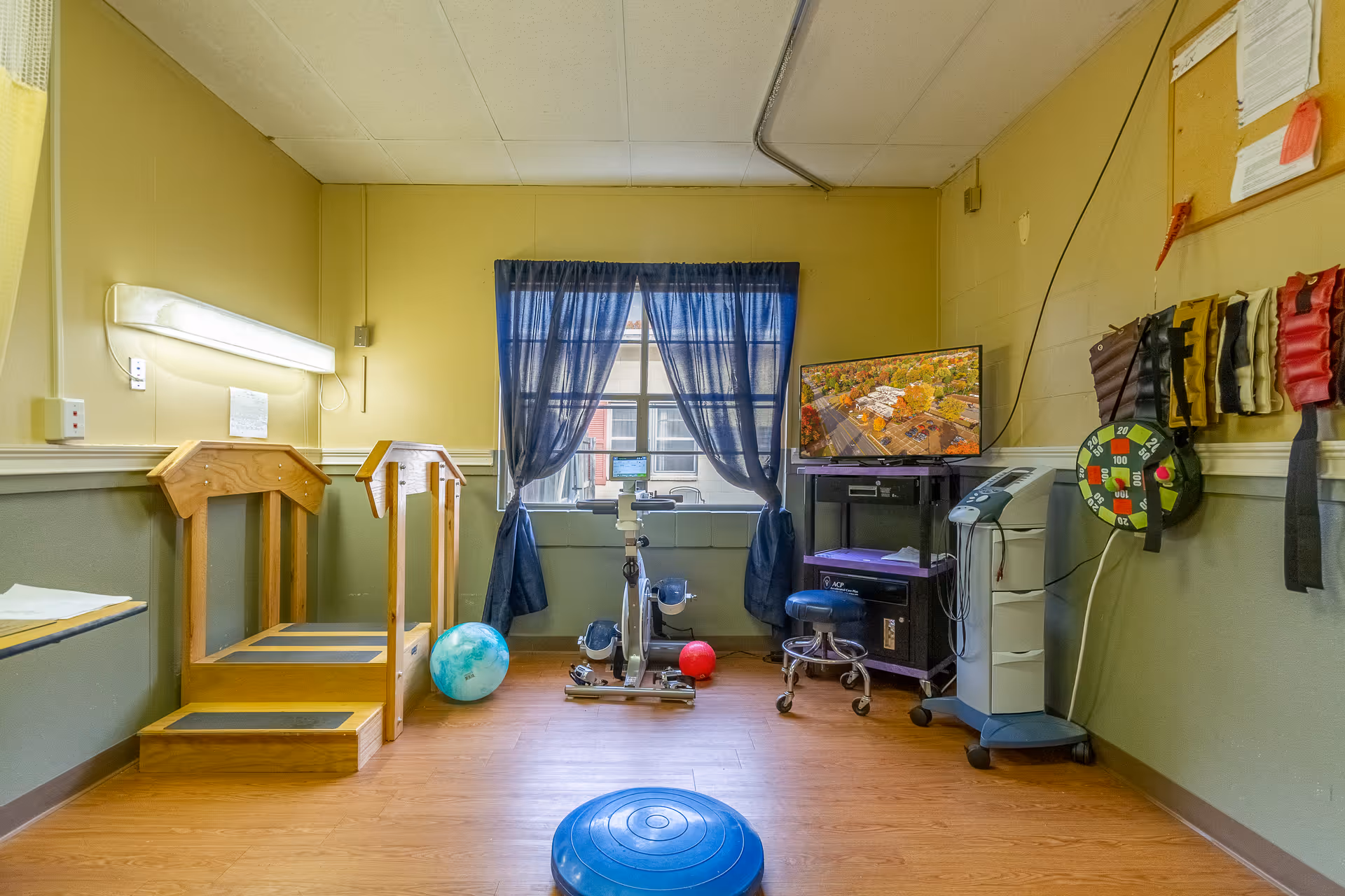 A small rehabilitation or therapy room with wooden steps for physical therapy, a stationary exercise bike, therapy balls, a balance cushion, a TV on a stand, and various therapy equipment mounted on the walls. The room has yellow walls, a window with blue curtains, and wood flooring.