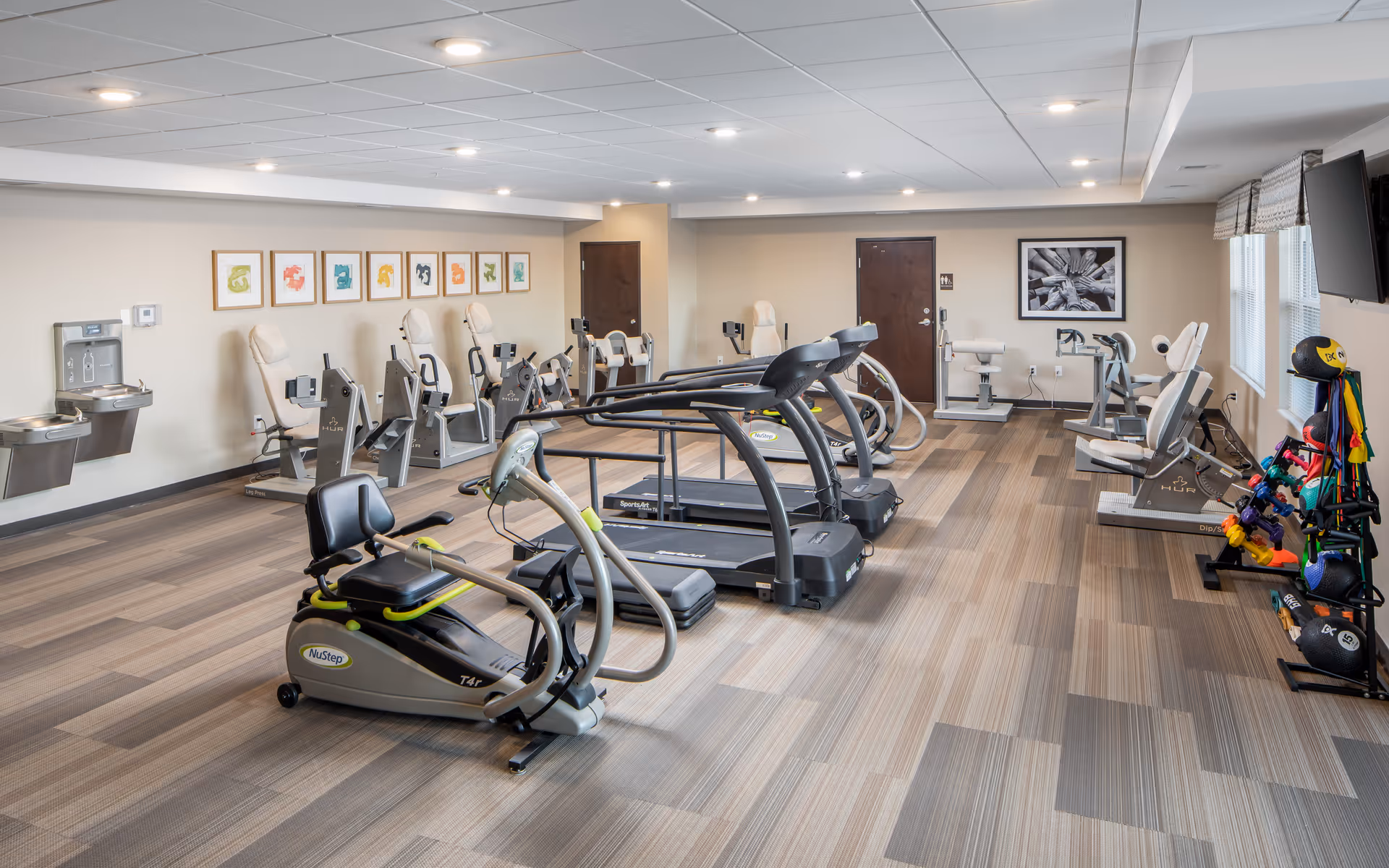 A spacious fitness room in a senior living facility featuring various exercise equipment including treadmills, stationary bikes, and strength training machines. The room has a carpeted floor, beige walls adorned with colorful framed artwork, and a rack with exercise balls and resistance bands near the windows. There are two water fountains on the left wall and a wall-mounted television on the right.