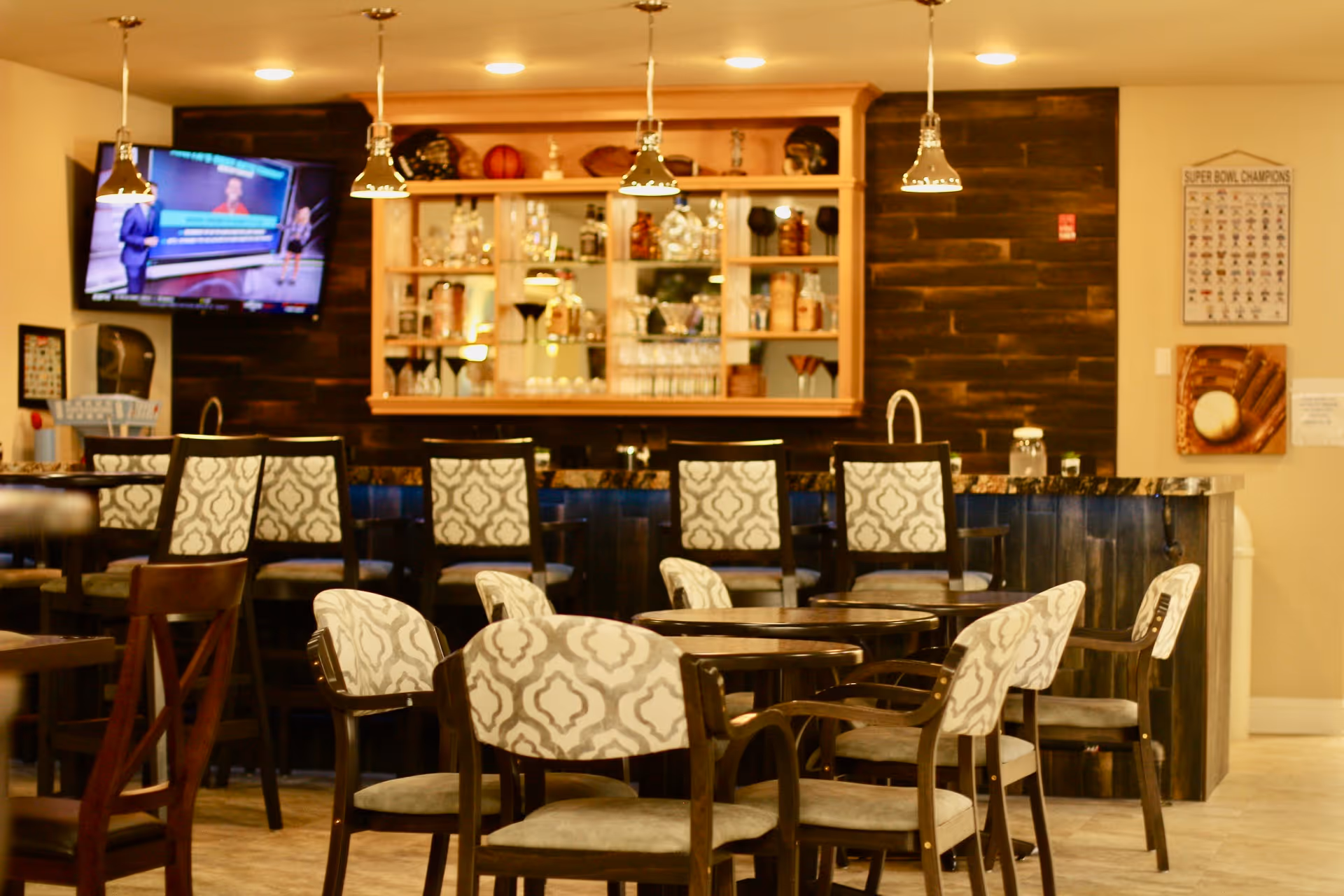 Well-lit dining and bar area with patterned chairs, round tables, and a TV mounted on the wall.
