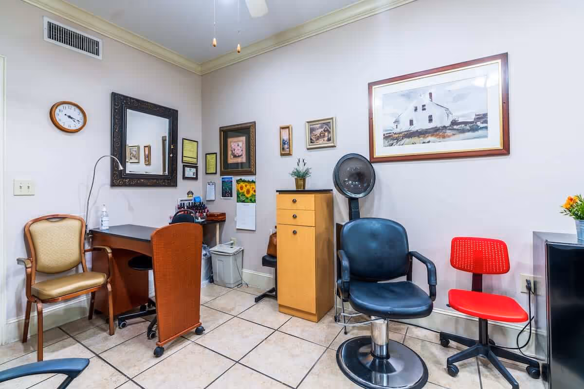 A small room with a black salon chair, a red rolling chair, a wooden cabinet, a desk with a brown chair, a beige chair, and various framed pictures and a clock on the walls. The floor is tiled and the walls are light-colored.