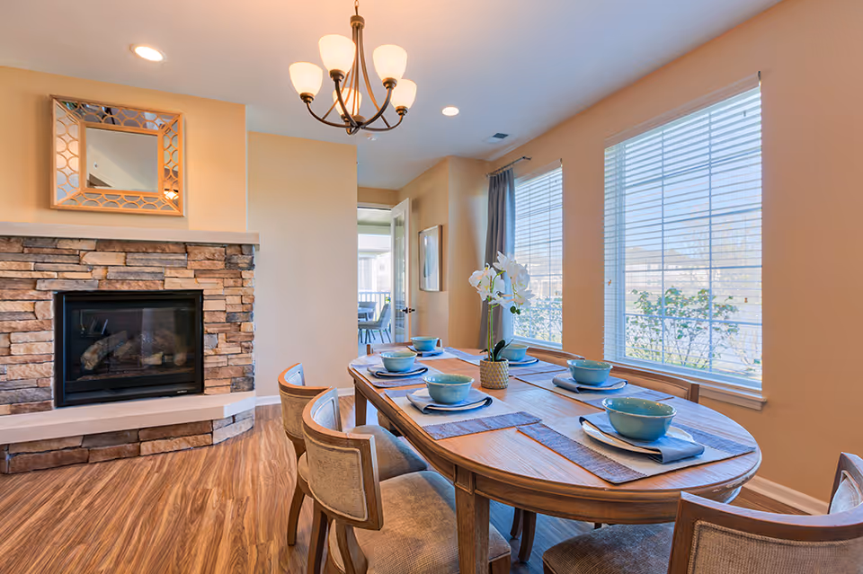 A cozy dining room with a wooden oval table set for six with blue bowls and napkins. The room features a stone fireplace with a decorative mirror above it, hardwood floors, and large windows with white blinds letting in natural light. A chandelier hangs above the table, and a door leads to another room in the background.