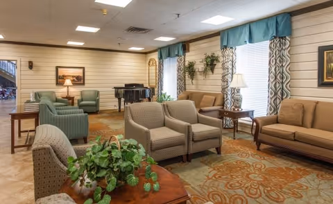 A cozy living room area in Winthrop Court featuring multiple upholstered armchairs and sofas arranged around wooden side tables. The room has large windows with patterned curtains and valances, a patterned carpet, wall-mounted plants, framed artwork, and a black grand piano in the corner.