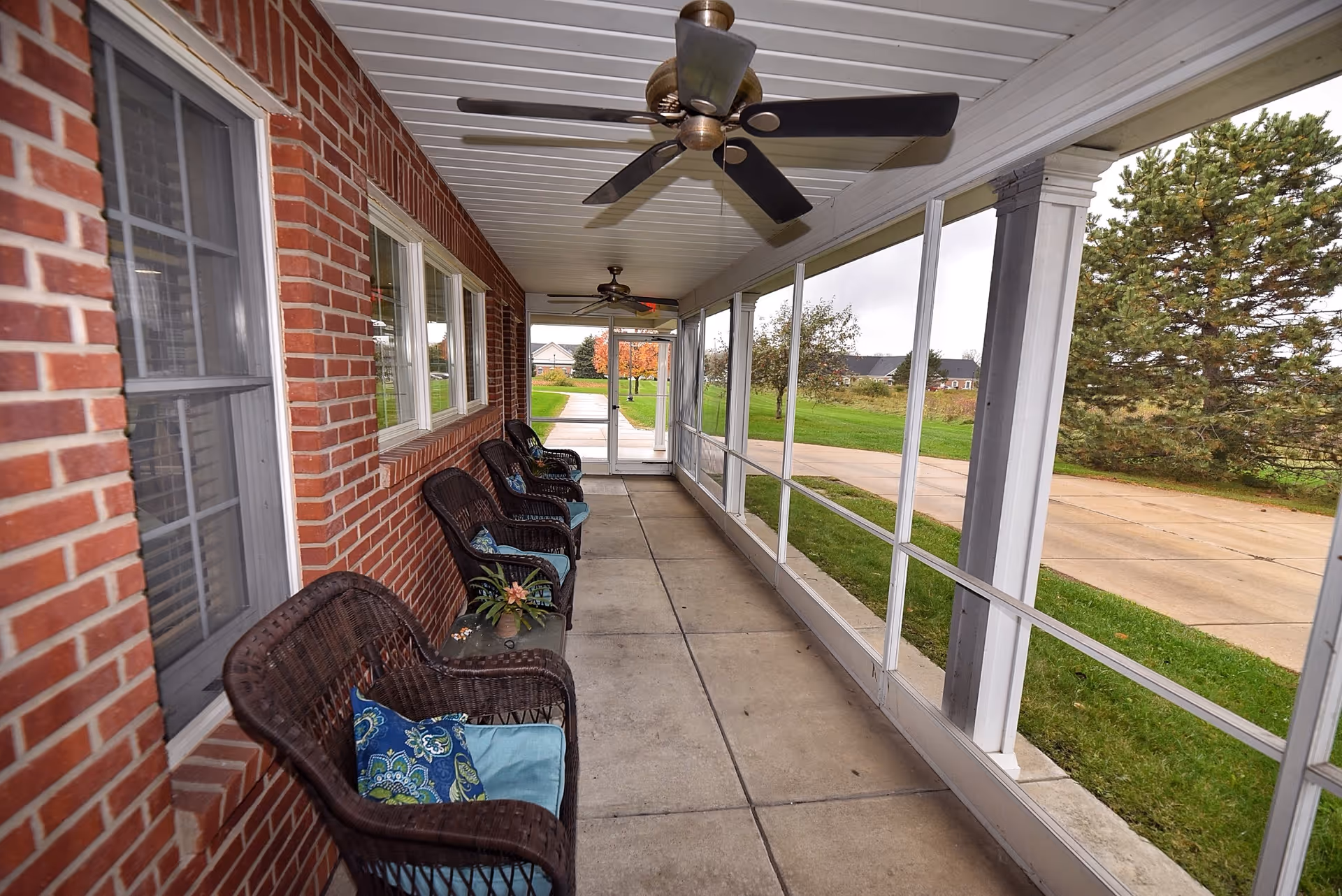 A covered outdoor porch area with a brick wall on one side and large screened windows on the other. The porch has several wicker chairs with cushions and pillows arranged along the brick wall. Ceiling fans are mounted on the white paneled ceiling. Outside the screened windows, there is a paved walkway, green grass, trees, and a cloudy sky.