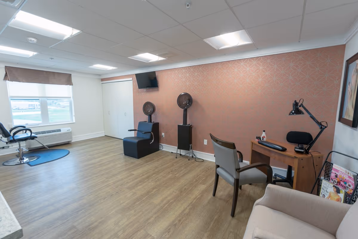 A bright and clean salon room in a senior living facility with wood flooring, a large window with a brown valance, two black hair dryer chairs against a patterned orange wall, a small TV mounted above them, a wooden desk with a black lamp and manicure supplies, a gray chair, a beige armchair, and a magazine rack filled with magazines.