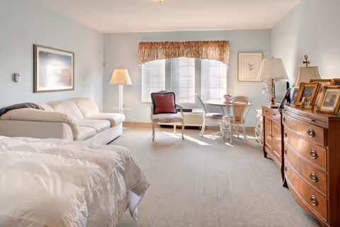 A bright and spacious bedroom with a large window letting in natural light. The room features a bed with white bedding, a white sofa, a wooden dresser with framed photos, a small round table with two chairs, and a floor lamp. The walls are painted light blue and decorated with framed artwork.