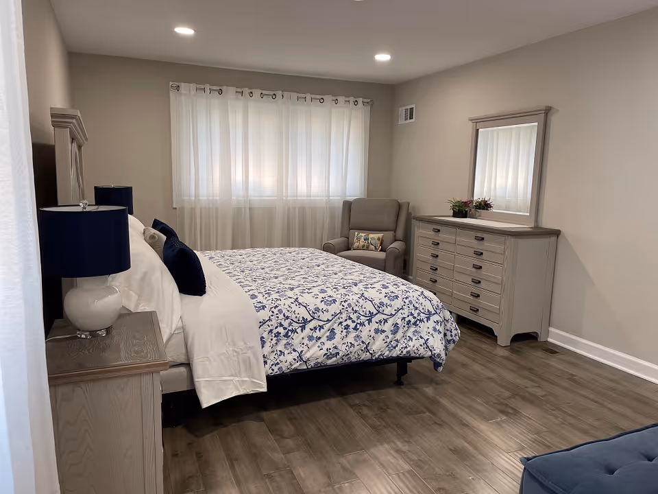 A bright, neatly furnished bedroom with a bed, nightstands, a dresser with mirror, and an armchair by a curtained window.