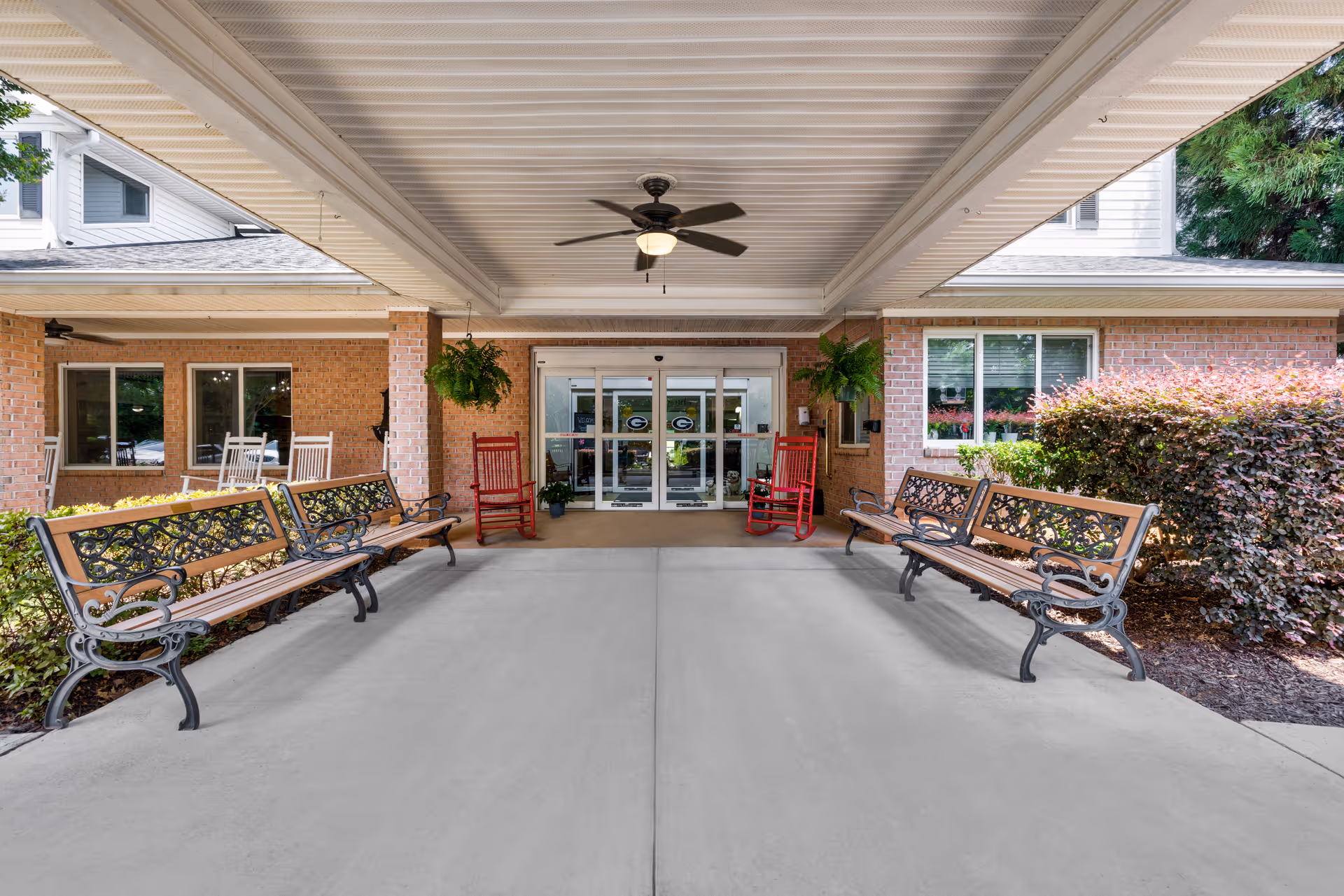 Covered entrance area of TerraBella Epps Bridge facility with two wooden benches with ornate metal armrests facing each other, two red rocking chairs near the glass automatic doors, hanging green plants, and a ceiling fan overhead. The building exterior is brick with windows and landscaping including bushes.