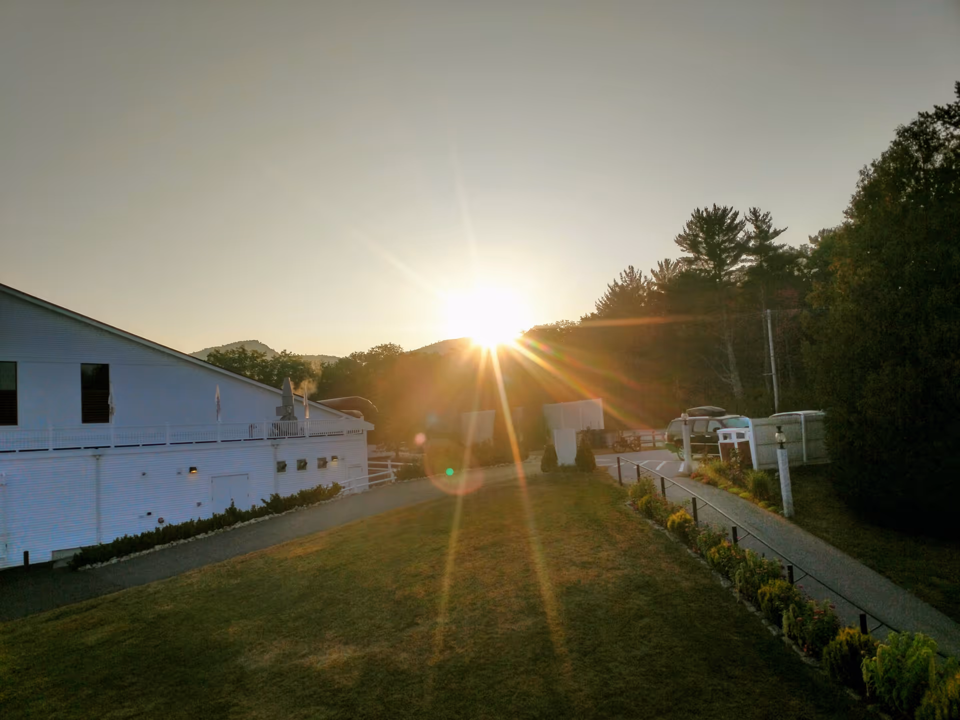 Sun setting behind trees and hills with a white building on the left and a pathway lined with plants on the right.