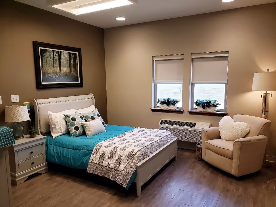 A cozy bedroom with a bed featuring a teal comforter and multiple pillows, a nightstand with a lamp and small plant, two windows with white blinds and flower pots on the windowsills, a beige armchair with a heart-shaped pillow, a floor lamp, and a framed picture of a forest on the wall.