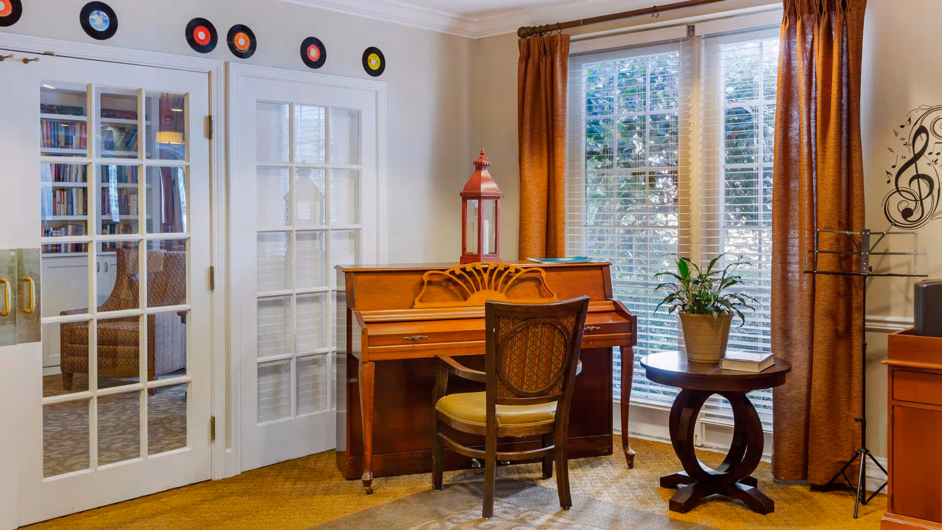 A cozy interior room featuring a wooden piano with a chair in front of it. On top of the piano is a red lantern and some books. To the right, there is a round wooden table with a potted plant and a book. Large windows with white blinds and brown curtains allow natural light to fill the room. The wall above the piano is decorated with vinyl records and a musical note decal. French doors with glass panels lead to another room with bookshelves and a chair.