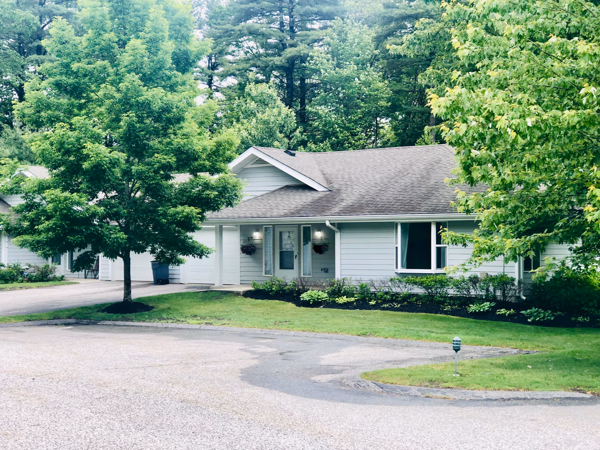 Single-story light-gray ranch-style senior living unit with a garage, front lawn, and surrounding trees.