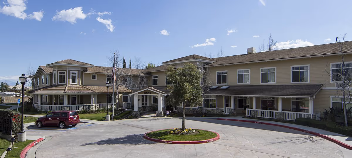 Exterior view of a two-story senior living facility building with beige siding and a covered entrance. There is a circular driveway with a small landscaped island in the center featuring a tree and flowers. A red SUV is parked near the entrance, and there are street lamps and a flagpole visible. The sky is clear with a few clouds.