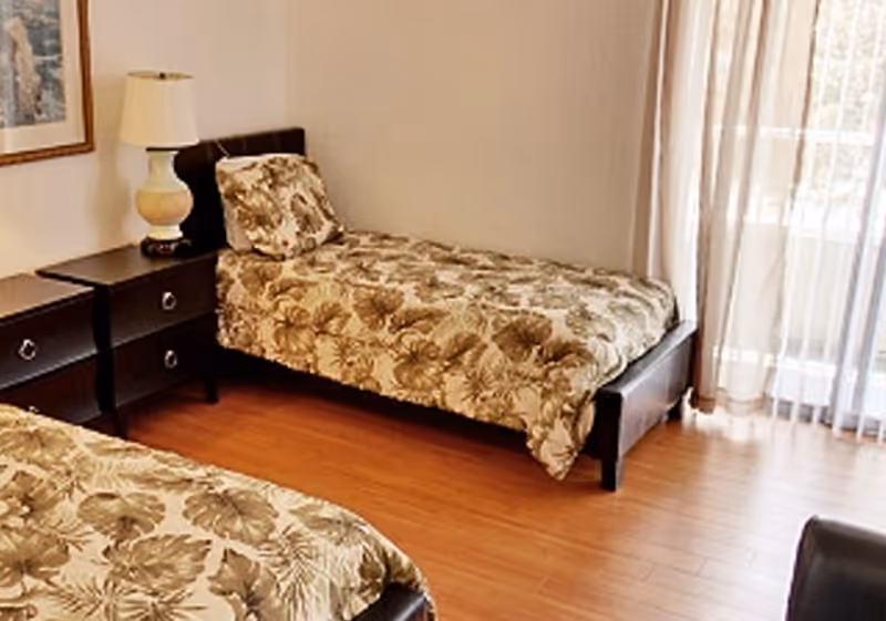 A bedroom with two single beds featuring matching floral bedspreads and pillows. There is a dark wooden nightstand with a lamp between the beds. The room has wooden flooring and a large window with sheer curtains allowing natural light to enter.
