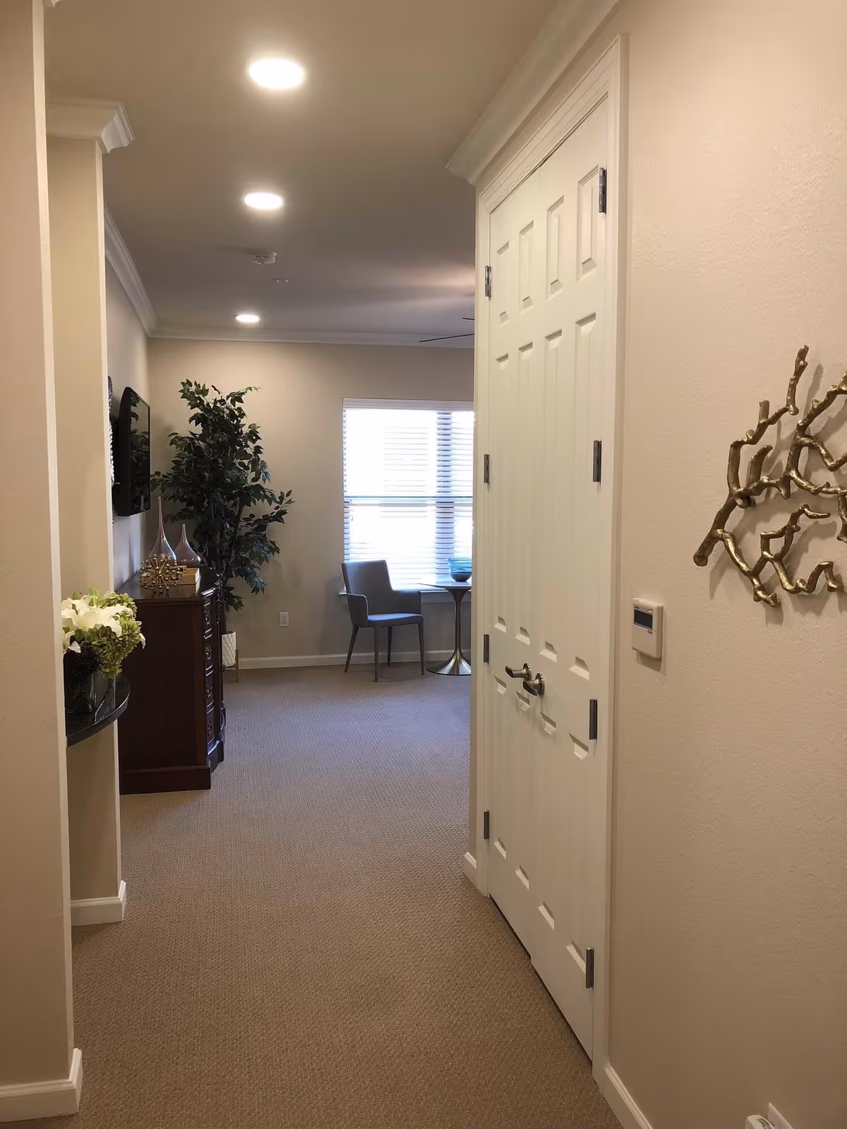 Interior hallway of a senior living facility with beige walls and carpet. On the right is a closed white double door and a gold decorative wall piece. At the end of the hallway is a window with blinds, a small round table, and two chairs. On the left side, there is a wooden cabinet with decorative items and a tall green plant next to it.