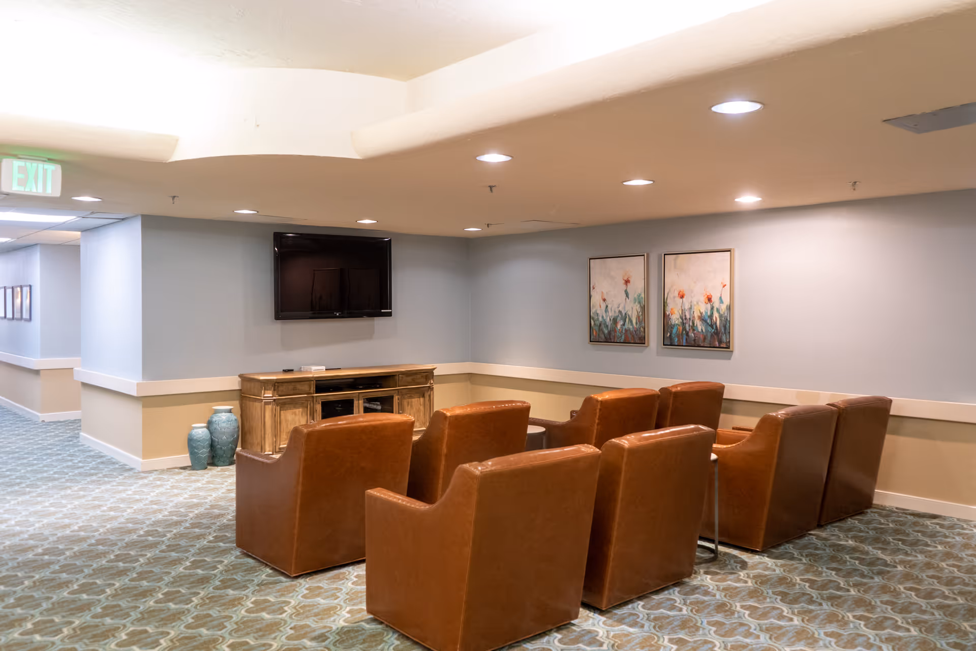 A cozy seating area with six brown leather armchairs arranged in two rows facing a wall-mounted flat screen TV. Below the TV is a wooden cabinet. The walls are painted light blue with a beige wainscoting, and two framed floral paintings hang on the wall to the right. The carpet has a patterned design, and recessed ceiling lights illuminate the space. There is an exit sign visible in the hallway to the left.