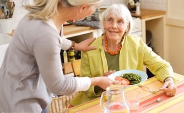 A caregiver hands a plate of food to an elderly woman seated at a dining table in a kitchen area.