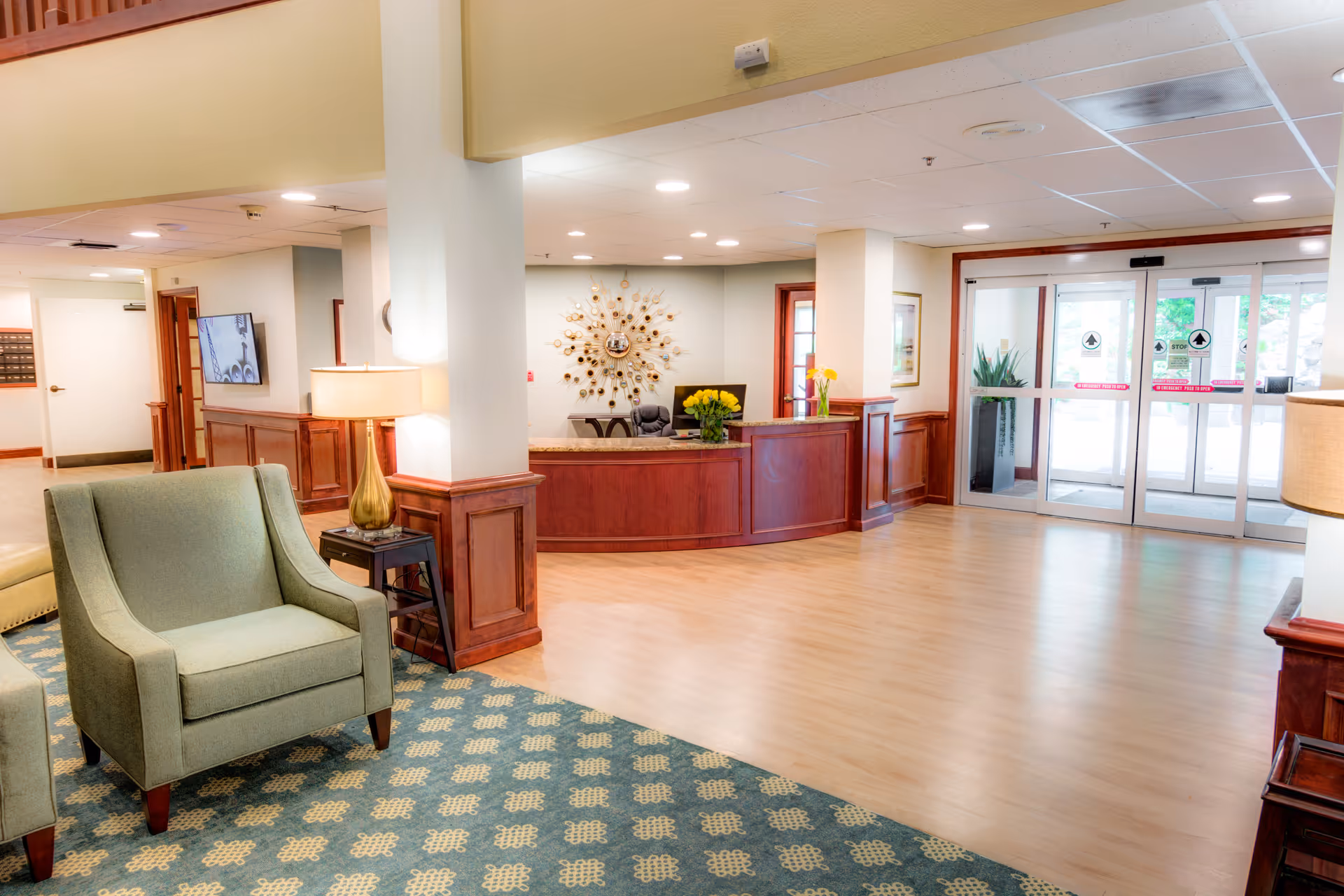 A bright and welcoming senior living facility lobby with a wooden reception desk adorned with yellow flowers, comfortable green armchairs on a patterned carpet, a side table with a lamp, and glass automatic entrance doors letting in natural light.