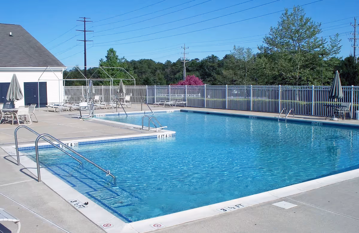 Outdoor swimming pool with clear blue water surrounded by a concrete deck. Several lounge chairs and tables with umbrellas are placed around the pool. A white fence encloses the pool area, and there are trees and greenery in the background under a clear blue sky.