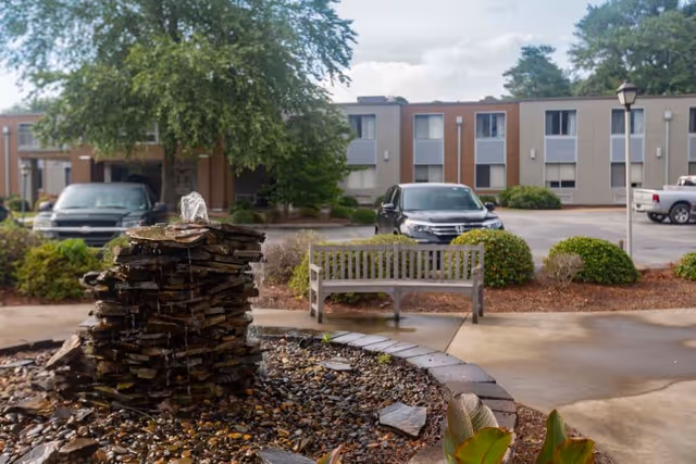 Outdoor view of Millennium Post Acute Rehabilitation facility showing a stone water fountain in the foreground, a wooden bench, parked cars, bushes, and a two-story building with multiple windows in the background under a partly cloudy sky.