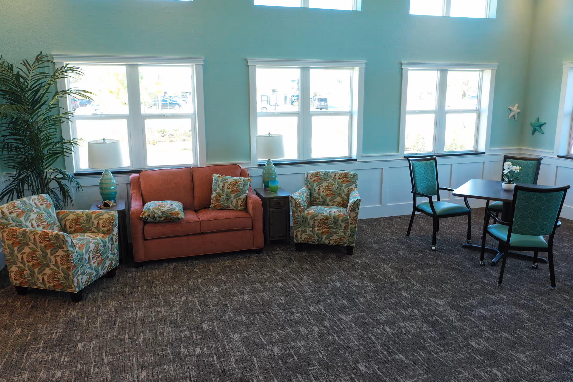 A bright and airy sitting area with large windows letting in natural light. The room features a coral-colored loveseat with patterned cushions, two matching armchairs with a tropical leaf design, a small side table with a lamp, and a square table with four teal cushioned chairs. The walls are painted light blue with white wainscoting, and there are decorative starfish on the wall.