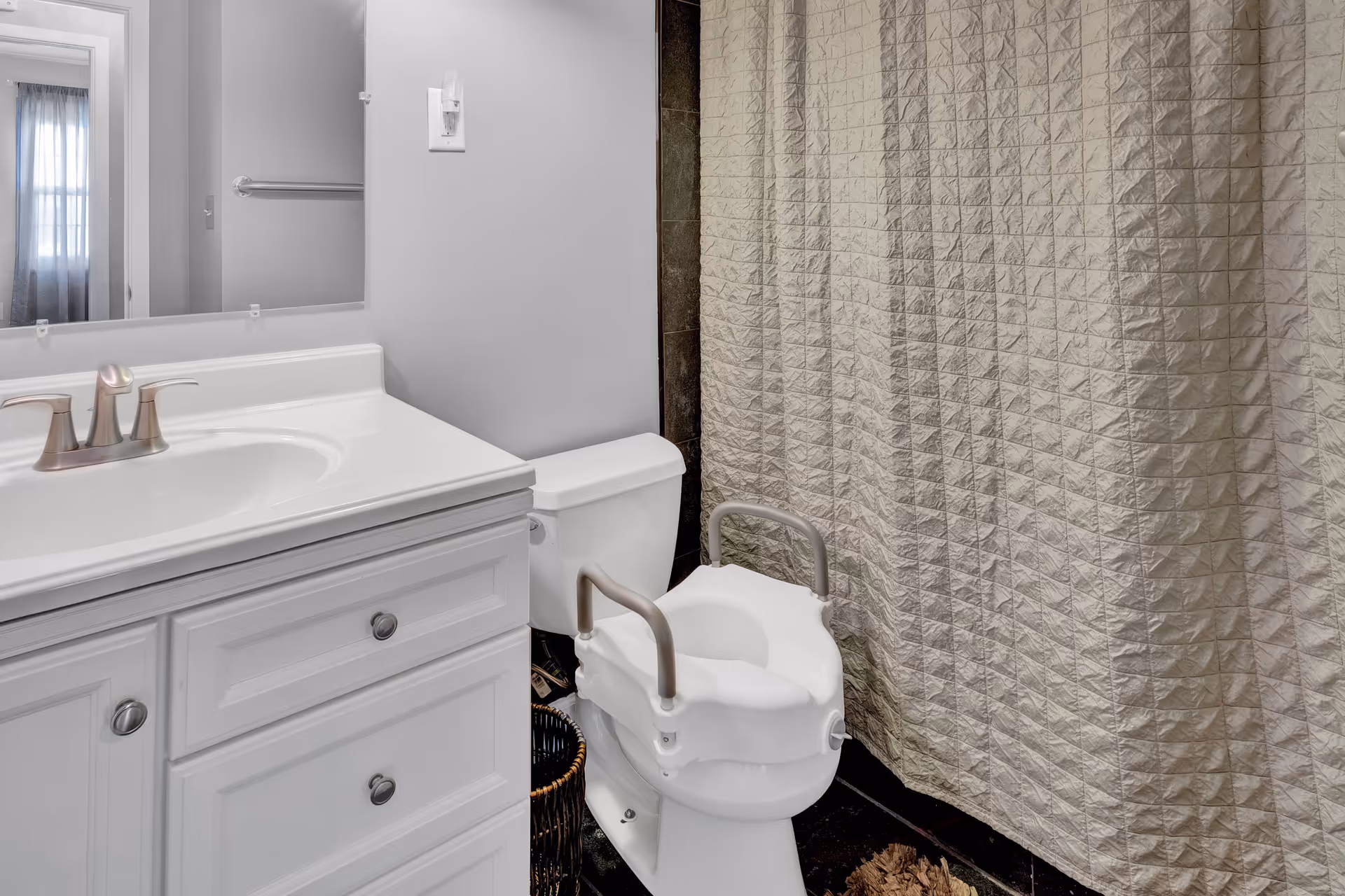 A bathroom with a white vanity and sink on the left, a toilet with a raised seat and armrests in the center, and a shower with a textured curtain on the right. A mirror above the vanity reflects a window with sheer curtains.