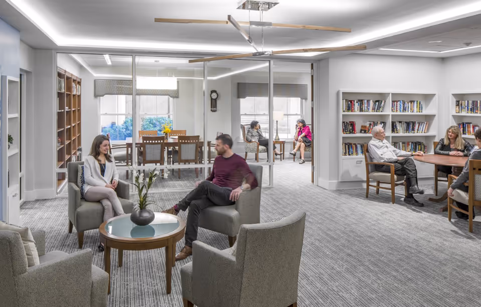 A bright community lounge in a senior living facility with armchairs, bookshelves, tables and several people talking and reading.
