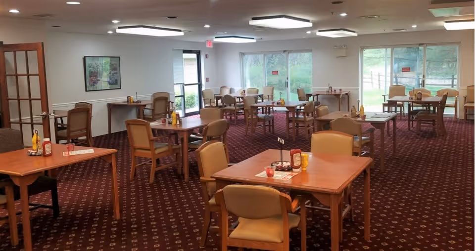 A dining room with multiple wooden tables and chairs arranged neatly on a patterned carpet. Each table has condiments such as ketchup and mustard bottles. Large windows and glass doors allow natural light to enter, showing greenery outside. The room has white walls with a framed picture and ceiling lights.